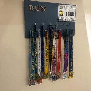 May include: A blue wooden board with the word "RUN" in gold letters. The board has hooks for hanging medals. There are several medals hanging on the hooks, including a gold medal with the text "Waterloo 10K Classic" and a purple medal with the text "5K Foam Fest".