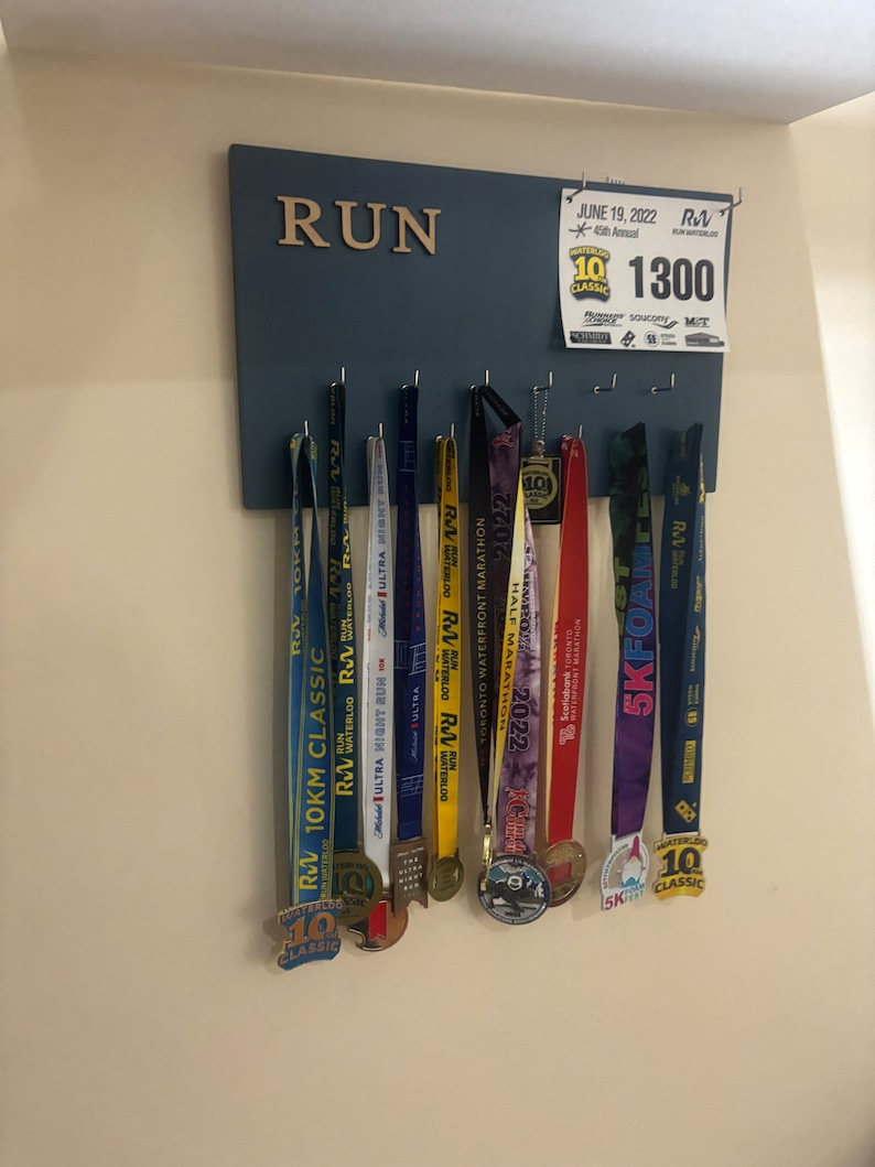 May include: A blue wooden board with the word "RUN" in gold letters. The board has hooks for hanging medals. There are several medals hanging on the hooks, including a gold medal with the text "Waterloo 10K Classic" and a purple medal with the text "5K Foam Fest".