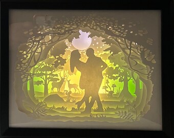 Puede incluir: Una obra de arte de papel recortado blanco que representa a una pareja bailando en un entorno de bosque. La pareja está silueteada contra una luna y estrellas brillantes. El bosque está detallado con árboles, ciervos y hongos.
