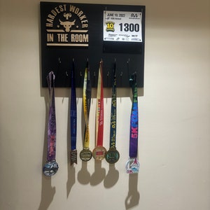 Puede incluir: Un tablero de madera negro con las palabras "Hardest Worker in the Room" en letras doradas. Un logotipo de cabeza de toro está por encima del texto. El tablero tiene ganchos para colgar medallas. Hay seis medallas colgadas en los ganchos, incluyendo una medalla morada con las palabras "5K Foam Fest" y una medalla roja, blanca y azul con las palabras "Waterloo Half Marathon".