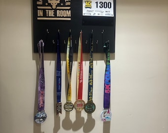 May include: A black wooden board with the words "Hardest Worker in the Room" in gold lettering. A bull's head logo is above the text. The board has hooks for hanging medals. There are six medals hanging on the hooks, including a purple medal with the words "5K Foam Fest" and a red, white, and blue medal with the words "Waterloo Half Marathon".