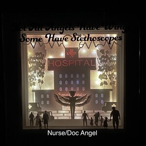 May include: A papercraft shadow box with a silhouette of an angel with wings, a hospital building, and a family. The text "Not All Angels Have Wings Some Have Stethoscopes" is at the top of the box. The text "HOSPITAL" is on the building. The text "Nurse/Doc Angel" is at the bottom of the box.