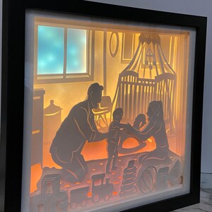 Op de afbeelding: Een 3D-schaduwdoos van papier met een verlichte sc&egrave;ne van een gezin in een kinderkamer. De vader knielt en houdt een baby vast, terwijl de moeder op de grond zit en naar de baby kijkt. Er liggen speelgoedjes op de grond, waaronder een schommelpaard, blokken en een speelgoedtrein.
