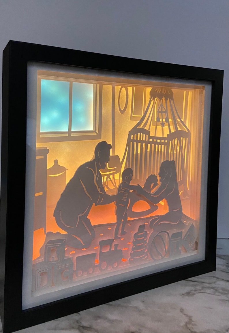 Op de afbeelding: Een 3D-schaduwdoos van papier met een verlichte sc&egrave;ne van een gezin in een kinderkamer. De vader knielt en houdt een baby vast, terwijl de moeder op de grond zit en naar de baby kijkt. Er liggen speelgoedjes op de grond, waaronder een schommelpaard, blokken en een speelgoedtrein.