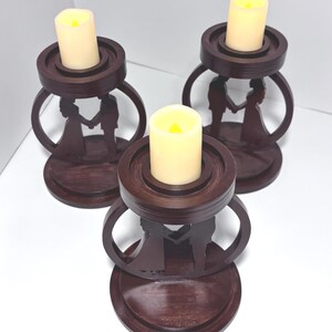 K&ouml;nnte beinhalten: Drei dunkelbraune Holzkerzenhalter mit einer Silhouette eines Paares, das sich an den H&auml;nden h&auml;lt. Jeder Halter hat einen runden Sockel, eine kreisf&ouml;rmige Oberseite und eine cremefarbene LED-Kerze.