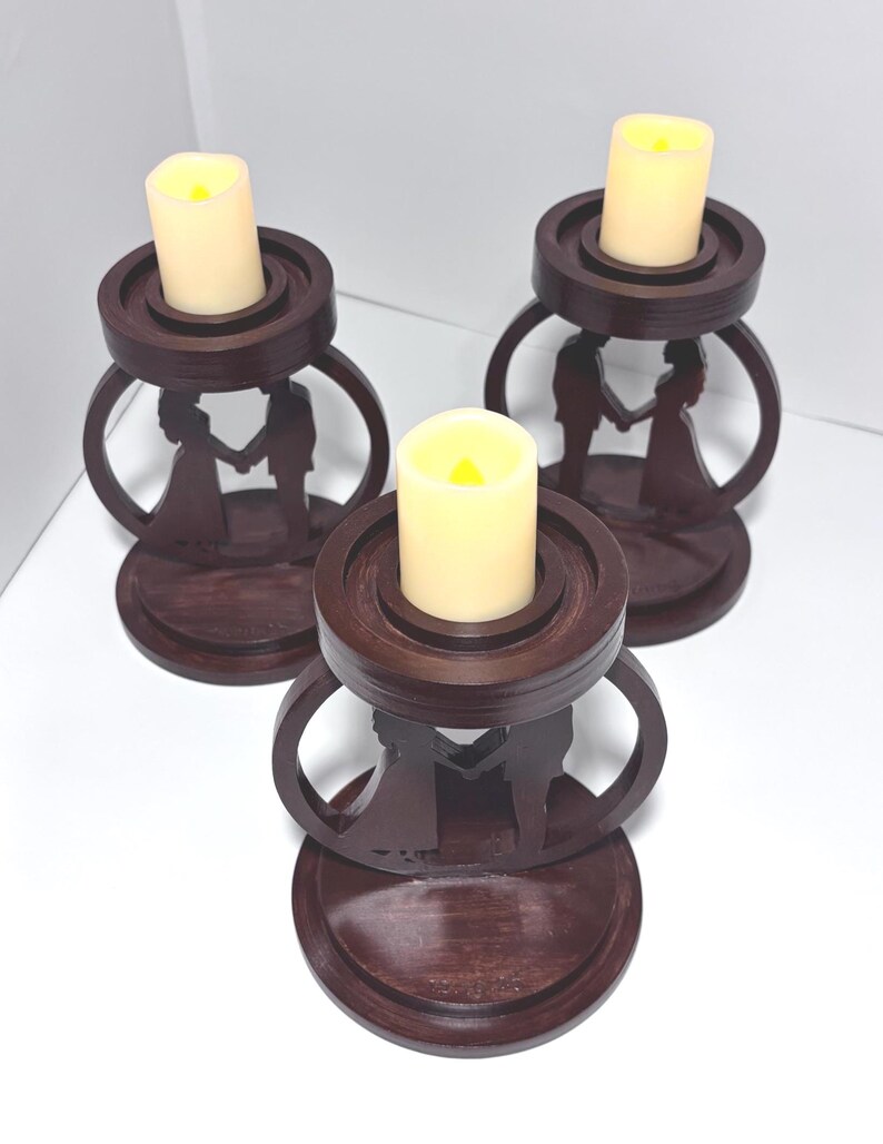 K&ouml;nnte beinhalten: Drei dunkelbraune Holzkerzenhalter mit einer Silhouette eines Paares, das sich an den H&auml;nden h&auml;lt. Jeder Halter hat einen runden Sockel, eine kreisf&ouml;rmige Oberseite und eine cremefarbene LED-Kerze.