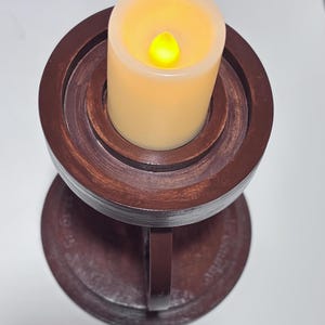 K&ouml;nnte beinhalten: Eine batteriebetriebene Kerze mit flackerndem gelbem Licht sitzt in einem dunkelbraunen Kerzenhalter aus Holz. Der Kerzenhalter hat einen runden Sockel und ein mehrstufiges Design mit einem Griff an der Seite.