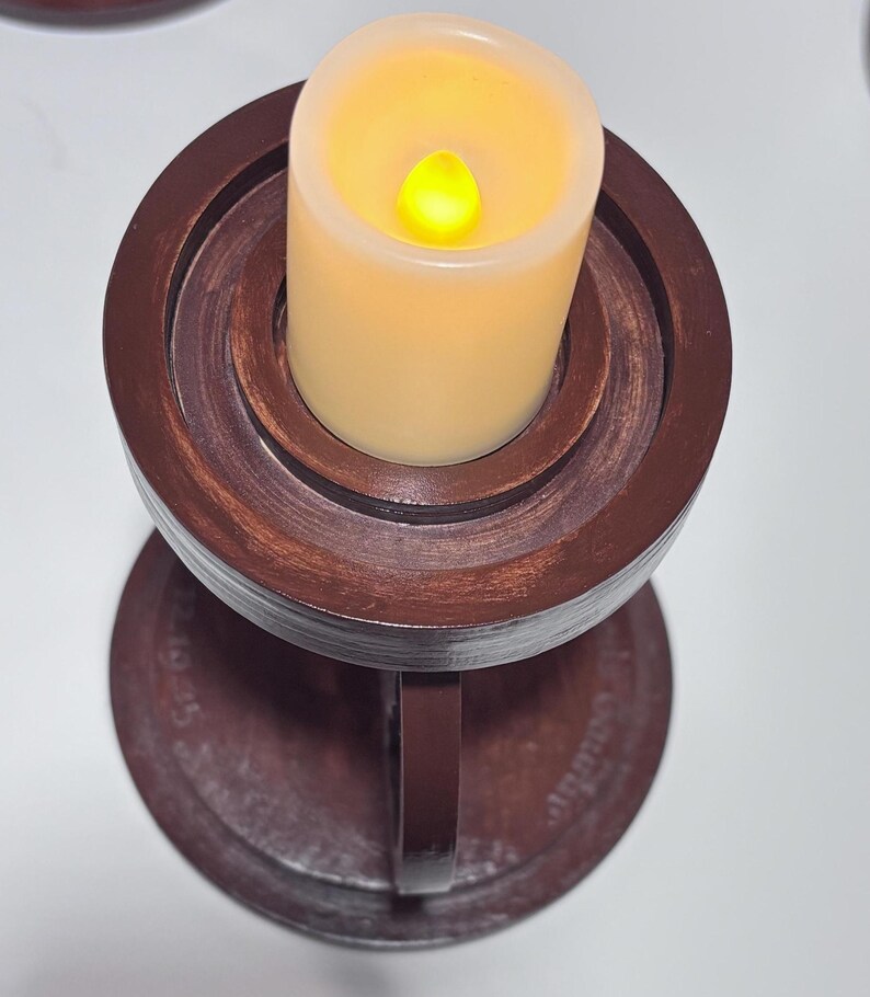 K&ouml;nnte beinhalten: Eine batteriebetriebene Kerze mit flackerndem gelbem Licht sitzt in einem dunkelbraunen Kerzenhalter aus Holz. Der Kerzenhalter hat einen runden Sockel und ein mehrstufiges Design mit einem Griff an der Seite.