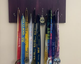 Puede incluir: Un soporte de madera para medallas con el texto "JUST DON'T QUIT" en letras marrones sobre un fondo morado. El soporte tiene ganchos para colgar medallas. Hay varias medallas de colores colgadas en los ganchos, incluyendo una medalla azul y amarilla con el texto "WATERLOO 10KM CLASSIC" y una medalla morada y blanca con el texto "HALF MARATHON".