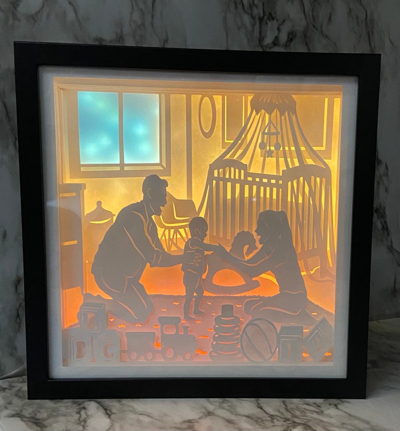 Op de afbeelding: Een 3D-schaduwdoos van papier met een familiesc&egrave;ne. De doos wordt van achteren verlicht, waardoor een warme gloed ontstaat. De sc&egrave;ne toont een gezin van drie, een vader, een moeder en een kind, die in een kamer met een wieg en speelgoed staan. De vader houdt de hand van het kind vast en de moeder kijkt naar het kind. De doos is gemaakt van wit papier en heeft een zwarte lijst.