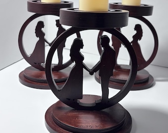 Puede incluir: Juego de tres candelabros de madera con acabado marrón oscuro. Cada uno presenta un diseño circular con la silueta de una pareja tomados de la mano. Los candelabros tienen una base redonda y una plataforma para una vela color crema.