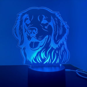 May include: A blue light illuminates a white silhouette of a dog's head, creating a 3D effect. The dog has floppy ears and a happy expression. The light is on a black base.