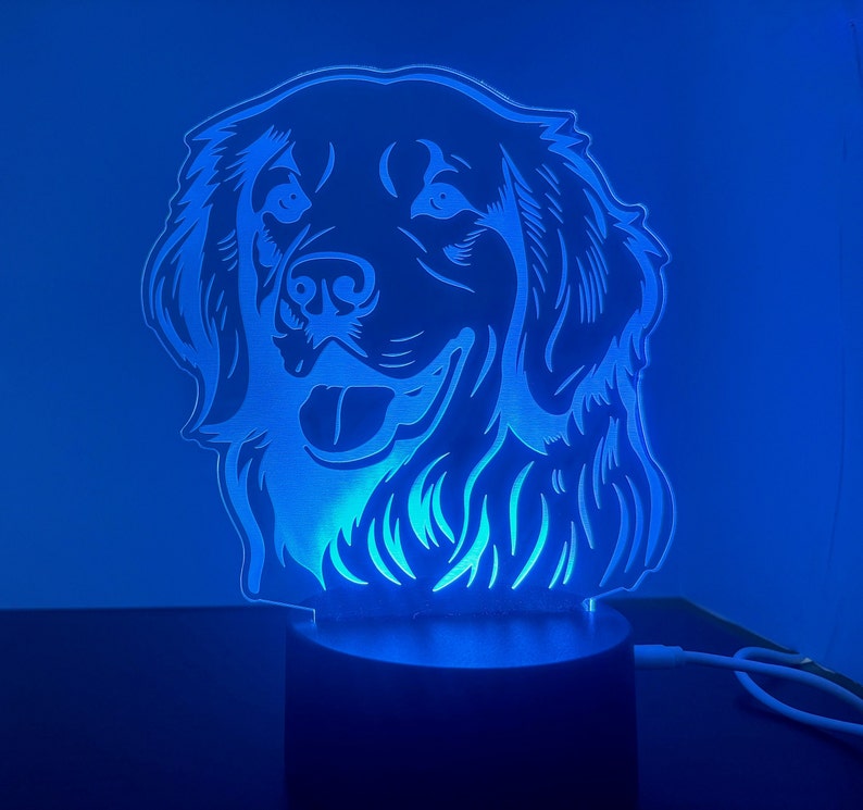 May include: A blue light illuminates a white silhouette of a dog's head, creating a 3D effect. The dog has floppy ears and a happy expression. The light is on a black base.