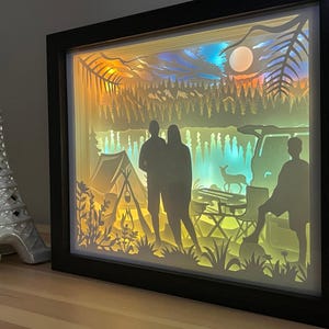 May include: A rectangular shadow box light featuring a layered paper cutout scene of a camping trip with silhouettes of three people, a tent, a vehicle, and a deer, illuminated by warm yellow and cool blue light, set in a black frame.