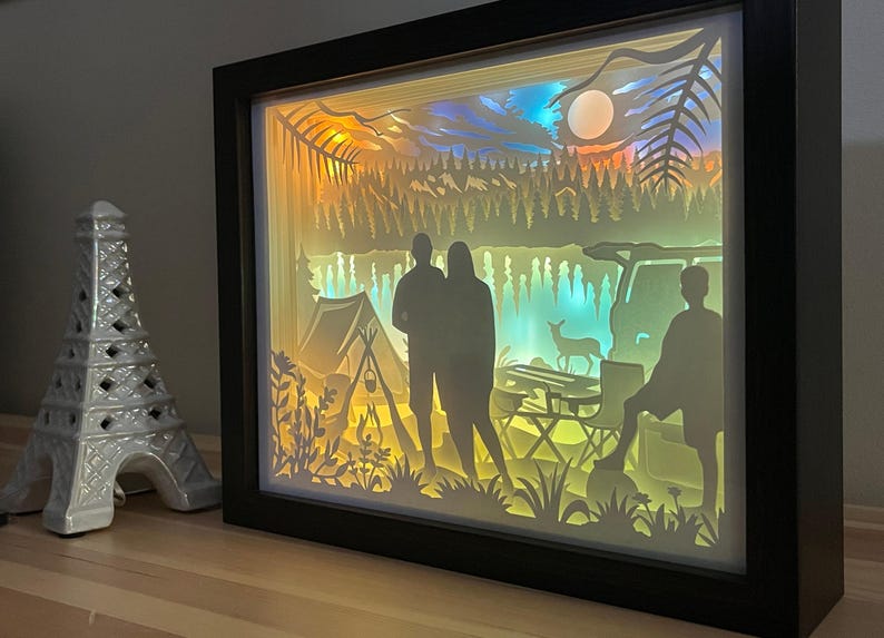 May include: A rectangular shadow box light featuring a layered paper cutout scene of a camping trip with silhouettes of three people, a tent, a vehicle, and a deer, illuminated by warm yellow and cool blue light, set in a black frame.