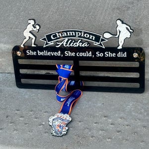 May include: Black acrylic medal display with a tennis player silhouette on each side. The display has a white inscription that reads "Champion Alisha She believed, She could, So She did". A blue and orange medal is hanging from the display.