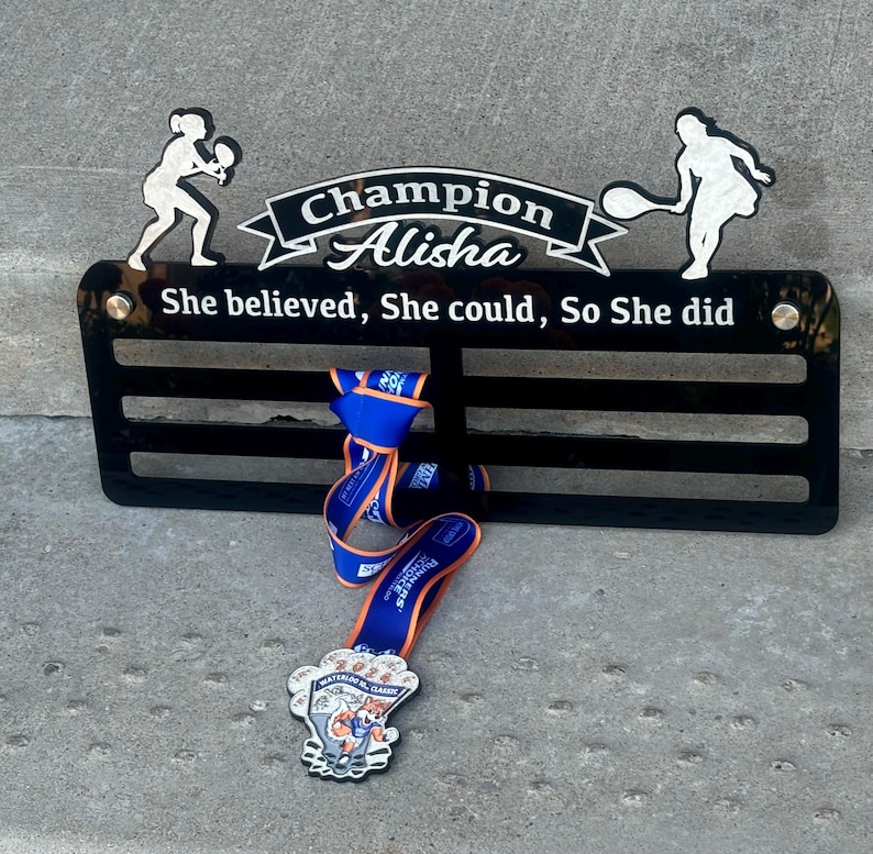 May include: Black acrylic medal display with a tennis player silhouette on each side. The display has a white inscription that reads "Champion Alisha She believed, She could, So She did". A blue and orange medal is hanging from the display.