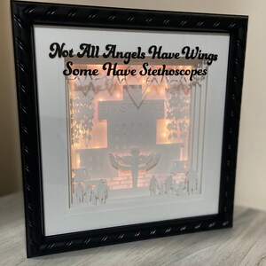 May include: A white framed shadow box with a 3D paper cut-out design of a hospital building. The design includes a cross, a hospital sign, trees, and a figure with wings. The text "Not All Angels Have Wings Some Have Stethoscopes" is written at the top of the frame.
