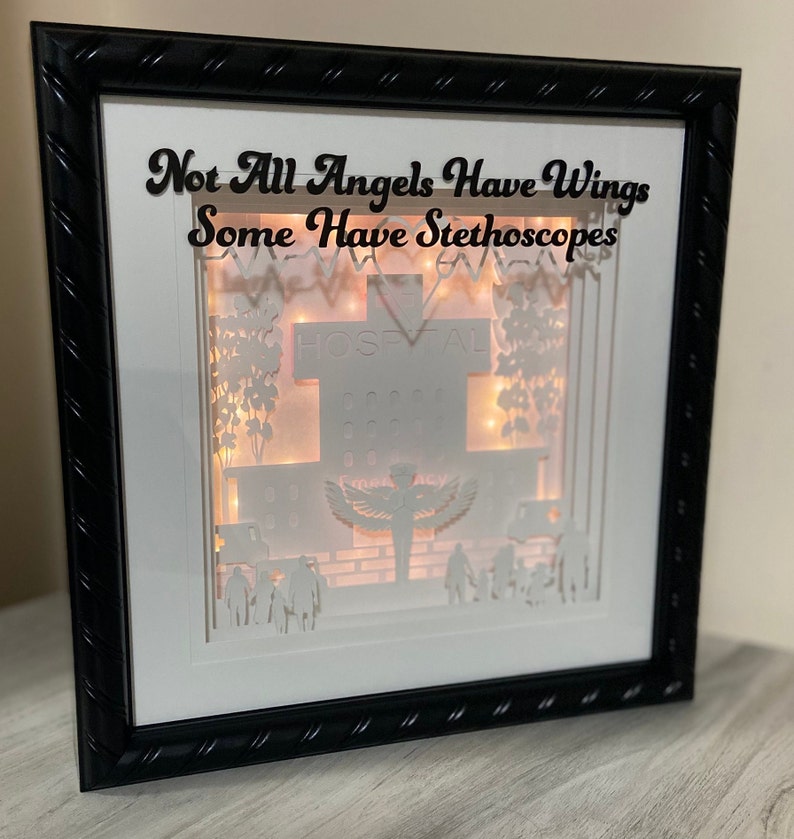 May include: A white framed shadow box with a 3D paper cut-out design of a hospital building. The design includes a cross, a hospital sign, trees, and a figure with wings. The text "Not All Angels Have Wings Some Have Stethoscopes" is written at the top of the frame.