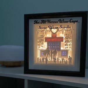May include: A black framed 3D paper art piece with a white background. The art features a hospital building with a red heart and the text "Not All Heroes Wear Capes, Some Wear Scrubs" and "Care with Love".