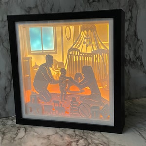 Op de afbeelding: Een 3D-schaduwdoos van papier met een gezinsscene. De sc&egrave;ne toont een gezin van drie, een vader, een moeder en een kind, in een kinderkamer. De vader staat, de moeder zit en het kind speelt met een schommelpaard. De kinderkamer heeft een wieg, een schommelpaard en speelgoed. De schaduwdoos wordt van achteren verlicht, waardoor een warme gloed ontstaat.
