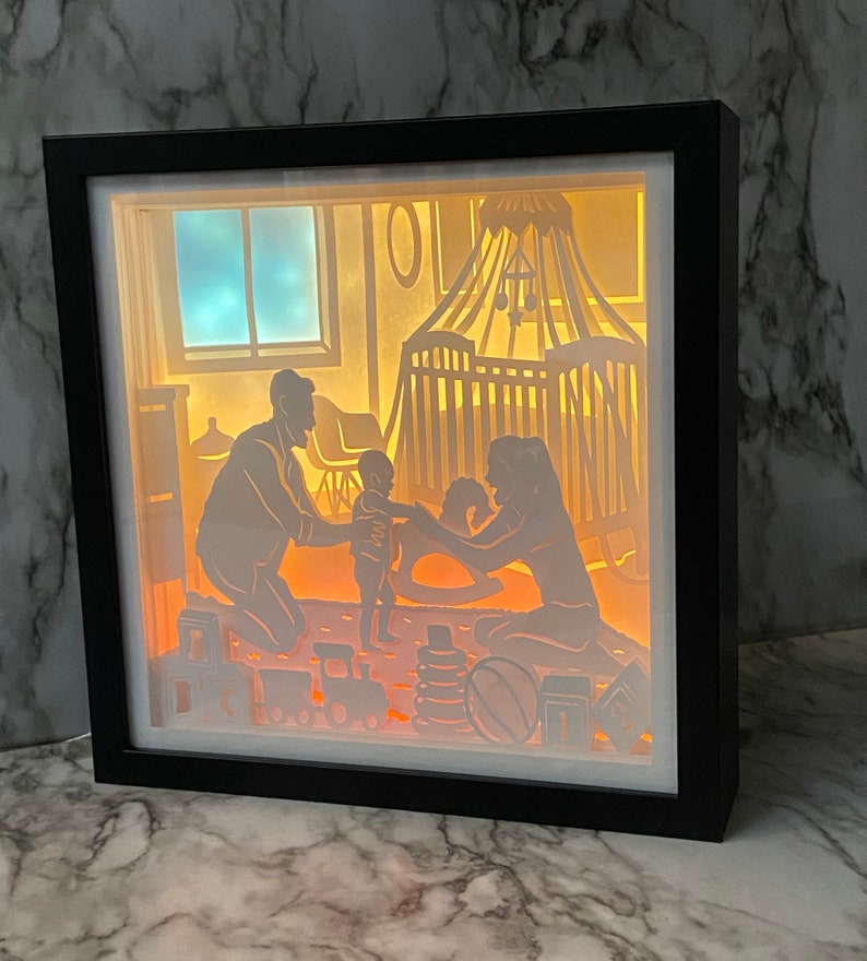 Op de afbeelding: Een 3D-schaduwdoos van papier met een gezinsscene. De sc&egrave;ne toont een gezin van drie, een vader, een moeder en een kind, in een kinderkamer. De vader staat, de moeder zit en het kind speelt met een schommelpaard. De kinderkamer heeft een wieg, een schommelpaard en speelgoed. De schaduwdoos wordt van achteren verlicht, waardoor een warme gloed ontstaat.