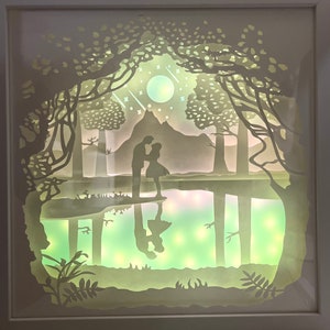 K&ouml;nnte beinhalten: Ein wei&szlig;es Papier-Schnittbild, das ein Paar zeigt, das in einem Wald an einem See steht. Die Szene ist mit einem sanften gr&uuml;nen Licht beleuchtet, das eine romantische Atmosph&auml;re schafft. Das Paar ist vor dem Licht silhouettiert, und die B&auml;ume und der See sind mit filigranen Papierschnitten detailliert dargestellt.