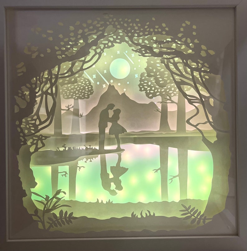 K&ouml;nnte beinhalten: Ein wei&szlig;es Papier-Schnittbild, das ein Paar zeigt, das in einem Wald an einem See steht. Die Szene ist mit einem sanften gr&uuml;nen Licht beleuchtet, das eine romantische Atmosph&auml;re schafft. Das Paar ist vor dem Licht silhouettiert, und die B&auml;ume und der See sind mit filigranen Papierschnitten detailliert dargestellt.