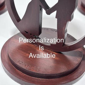 K&ouml;nnte beinhalten: Ein dunkelbrauner Kuchenaufsatz f&uuml;r eine Hochzeit mit einer Braut und einem Br&auml;utigam, die sich an den H&auml;nden halten, innerhalb eines kreisf&ouml;rmigen Designs. Der Text "Personalisierung ist verf&uuml;gbar" ist sichtbar. Das Datum "12.19.25" ist ebenfalls auf der Basis eingraviert.
