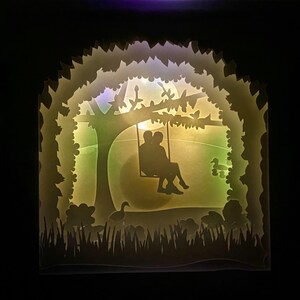 K&ouml;nnte beinhalten: Eine wei&szlig;e Papierschnitt-Silhouette eines Paares, das auf einer Schaukel unter einem Baum sitzt, mit einem Licht, das durch das Papier scheint. Der Hintergrund ist ein gr&uuml;nes Feld mit ein paar kleinen Blumen und V&ouml;geln.