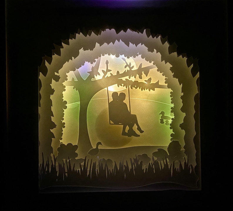 K&ouml;nnte beinhalten: Eine wei&szlig;e Papierschnitt-Silhouette eines Paares, das auf einer Schaukel unter einem Baum sitzt, mit einem Licht, das durch das Papier scheint. Der Hintergrund ist ein gr&uuml;nes Feld mit ein paar kleinen Blumen und V&ouml;geln.