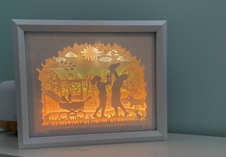 K&ouml;nnte beinhalten: Ein wei&szlig;er Rahmen mit einem Licht im Inneren, der eine Papierschnitt-Silhouette einer Familie mit einem Baby im Kinderwagen, einem Hund und einer Sonne im Hintergrund zeigt.