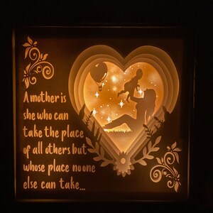 K&ouml;nnte beinhalten: Eine Holz-Schattenbox mit einem 3D-Papier-Ausschnitt-Design einer Mutter, die ein Kind in Herzform h&auml;lt. Die Box wird von hinten beleuchtet und erzeugt so ein warmes Leuchten. Der Spruch "Eine Mutter ist diejenige, die den Platz aller anderen einnehmen kann, aber deren Platz niemand sonst einnehmen kann..." ist auf der linken Seite der Box aufgedruckt.
