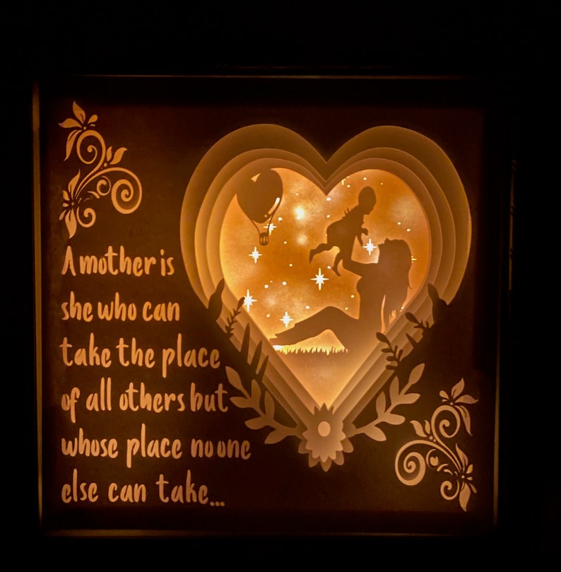 K&ouml;nnte beinhalten: Eine Holz-Schattenbox mit einem 3D-Papier-Ausschnitt-Design einer Mutter, die ein Kind in Herzform h&auml;lt. Die Box wird von hinten beleuchtet und erzeugt so ein warmes Leuchten. Der Spruch "Eine Mutter ist diejenige, die den Platz aller anderen einnehmen kann, aber deren Platz niemand sonst einnehmen kann..." ist auf der linken Seite der Box aufgedruckt.