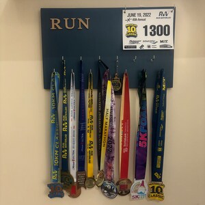 May include: A blue wooden board with the word "RUN" in gold letters. The board has hooks for hanging medals. There are 9 medals hanging on the hooks, all with different designs and colours. The medals are from various races, including a 10km classic, a half marathon, and a 5k foam fest. A sign above the board reads "JUNE 19, 2022, 45th Annual Waterloo 10 Classic, 1300".