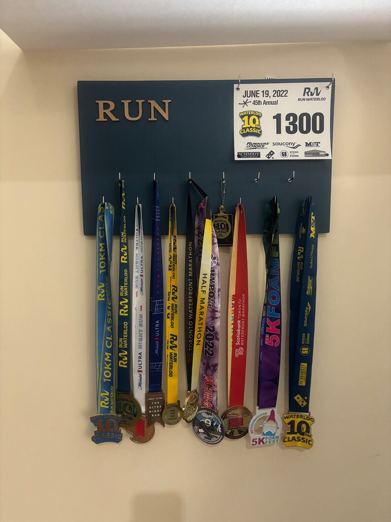 May include: A blue wooden board with the word "RUN" in gold letters. The board has hooks for hanging medals. There are 9 medals hanging on the hooks, all with different designs and colours. The medals are from various races, including a 10km classic, a half marathon, and a 5k foam fest. A sign above the board reads "JUNE 19, 2022, 45th Annual Waterloo 10 Classic, 1300".