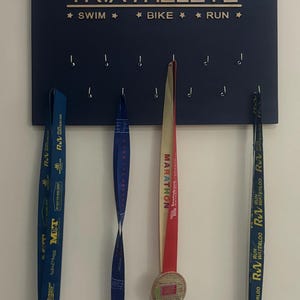 K&ouml;nnte beinhalten: Ein dunkelblaues Holzbrett mit den Worten "TRIATHELETE", "SWIM", "BIKE" und "RUN" in goldenen Buchstaben. Das Brett hat Haken zum Aufh&auml;ngen von Medaillen. Drei Medaillen h&auml;ngen an den Haken. Die Medaillen sind f&uuml;r einen 10-km-Lauf, einen Marathon und einen Nachtlauf.