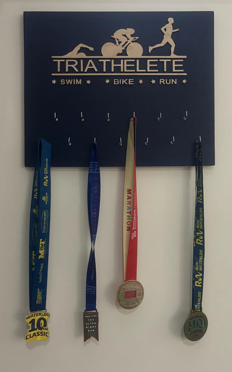 K&ouml;nnte beinhalten: Ein dunkelblaues Holzbrett mit den Worten "TRIATHELETE", "SWIM", "BIKE" und "RUN" in goldenen Buchstaben. Das Brett hat Haken zum Aufh&auml;ngen von Medaillen. Drei Medaillen h&auml;ngen an den Haken. Die Medaillen sind f&uuml;r einen 10-km-Lauf, einen Marathon und einen Nachtlauf.