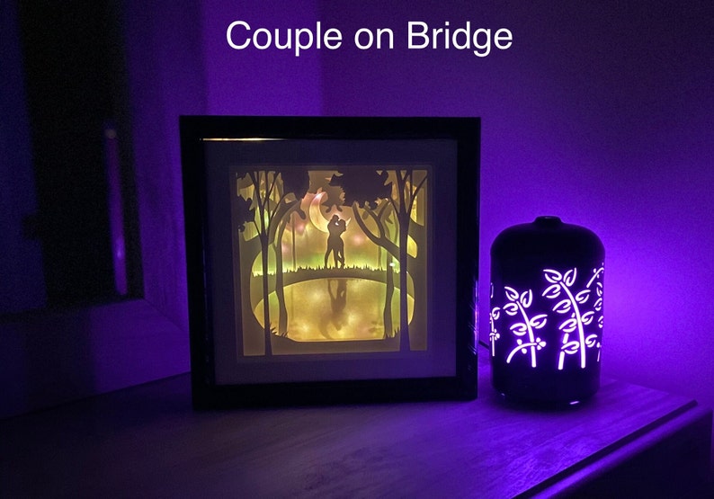 May include: A framed paper cut-out art piece depicting a couple silhouetted against a moonlit forest scene. The couple are standing on a bridge over a pond. The art piece is illuminated with warm white LED lights. A black diffuser with a white floral design is in the background.