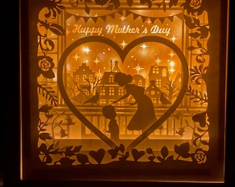 Puede incluir: Una obra de arte de papel recortado blanco con un marco en forma de corazón. El marco está decorado con un diseño floral y las palabras "Feliz Día de la Madre". Dentro del marco, una madre y un niño se representan en silueta, de pie frente a una ciudad.