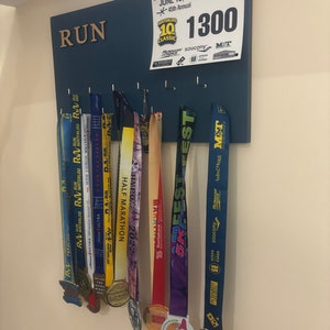 May include: A blue wooden board with the word "RUN" in gold letters. The board has hooks for hanging medals. There are several medals hanging on the hooks, including a blue and yellow medal with the text "WATERLOO 10K CLASSIC" and a blue and white medal with the text "5K FEST".
