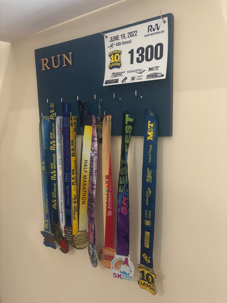 May include: A blue wooden board with the word "RUN" in gold letters. The board has hooks for hanging medals. There are several medals hanging on the hooks, including a blue and yellow medal with the text "WATERLOO 10K CLASSIC" and a blue and white medal with the text "5K FEST".