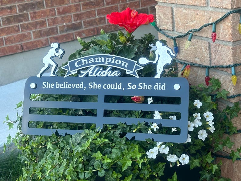 May include: Black metal plaque with a white silhouette of a tennis player, the text "Champion Alisha" and the quote "She believed, She could, So She did".