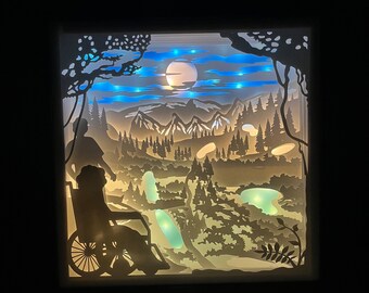 Puede incluir: Una obra de arte de papel recortado que representa a una persona en silla de ruedas con un paisaje de montaña en el fondo. La escena está iluminada con luces azules y blancas, creando un ambiente tranquilo y sereno.