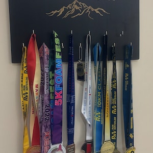 Puede incluir: Un tablero de madera negro con el texto "The Mountain Is You" en letras doradas. El tablero tiene ganchos para colgar medallas. Hay varias medallas de colores colgadas en los ganchos, incluidas medallas del "Waterloo Marathon", "5K Foam Fest" y "10km Classic".