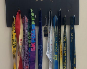May include: A black wooden board with the text "The Mountain Is You" in gold lettering. The board has hooks for hanging medals. There are several colourful medals hanging on the hooks, including medals from the "Waterloo Marathon", "5K Foam Fest", and "10km Classic".