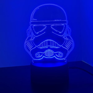 以下が含まれることがあります： スターウォーズのストームトルーパーのヘルメットの形をした青いLEDナイトライト。ライトは点灯しており、ヘルメットを照らしています。