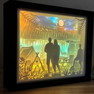 May include: A rectangular shadow box with a black frame featuring a multi-layered paper cutout scene of a camping trip. The backlit display shows silhouettes of two adults and a child, a tent, a campfire, a vehicle, a deer, and pine trees.