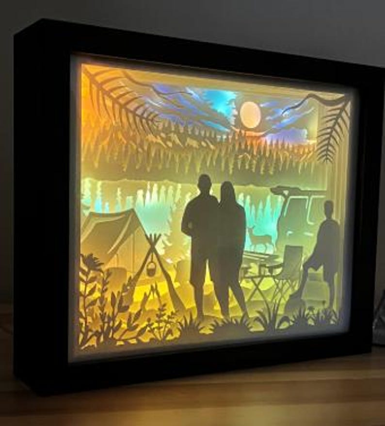 May include: A rectangular shadow box with a black frame featuring a multi-layered paper cutout scene of a camping trip. The backlit display shows silhouettes of two adults and a child, a tent, a campfire, a vehicle, a deer, and pine trees.