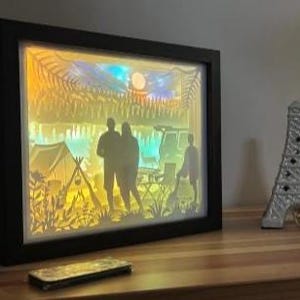 May include: A rectangular black shadow box frame containing a multi-layered paper cutout scene of a camping trip. The backlit display features silhouettes of two people standing near a tent, a vehicle, and trees under a moonlit sky with warm yellow and blue lighting.